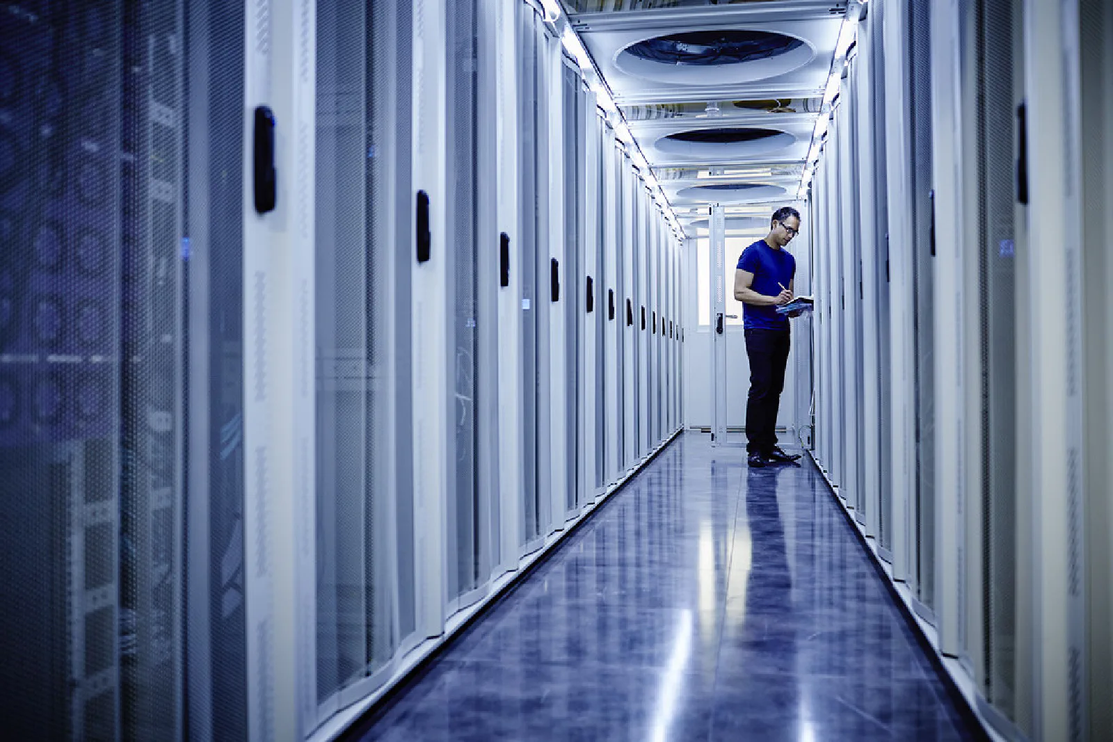 A lone technician with a tablet in a long, brightly lit data centre server aisle